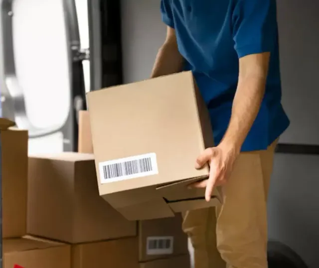 Profissional de uniforme azul carregando uma caixa de papelão com etiqueta de código de barras na porta do baú do caminhão